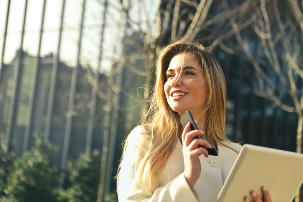 Smiling female real estate agent standing outdoors, holding a smartphone and notepad