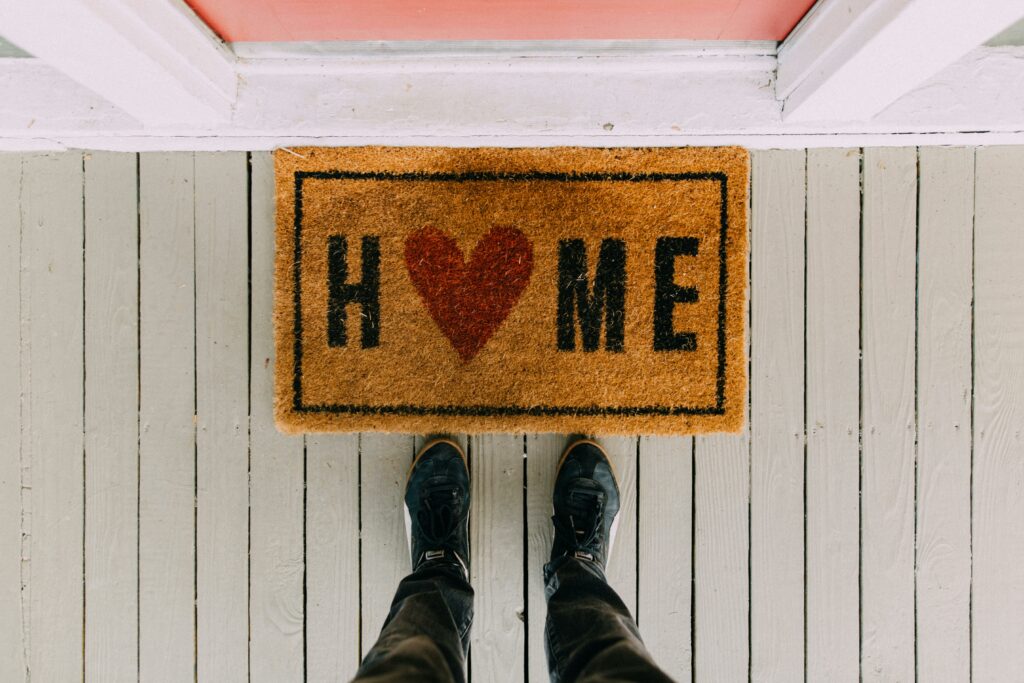 A Realtor stands at a front door delivering a hanger for local real estate marketing, with a welcome mat that says ‘home’ on it